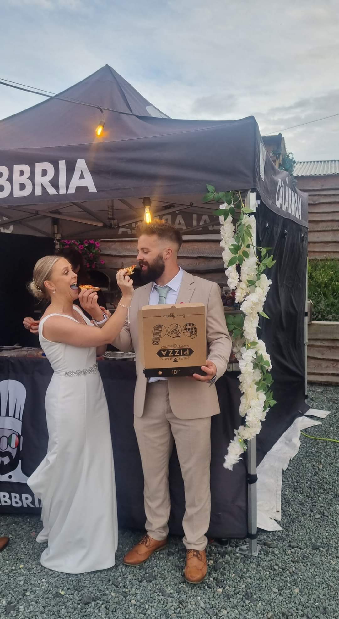 Bride and groom laughing together, feeding each other pizza in front of the Calàbbria stall