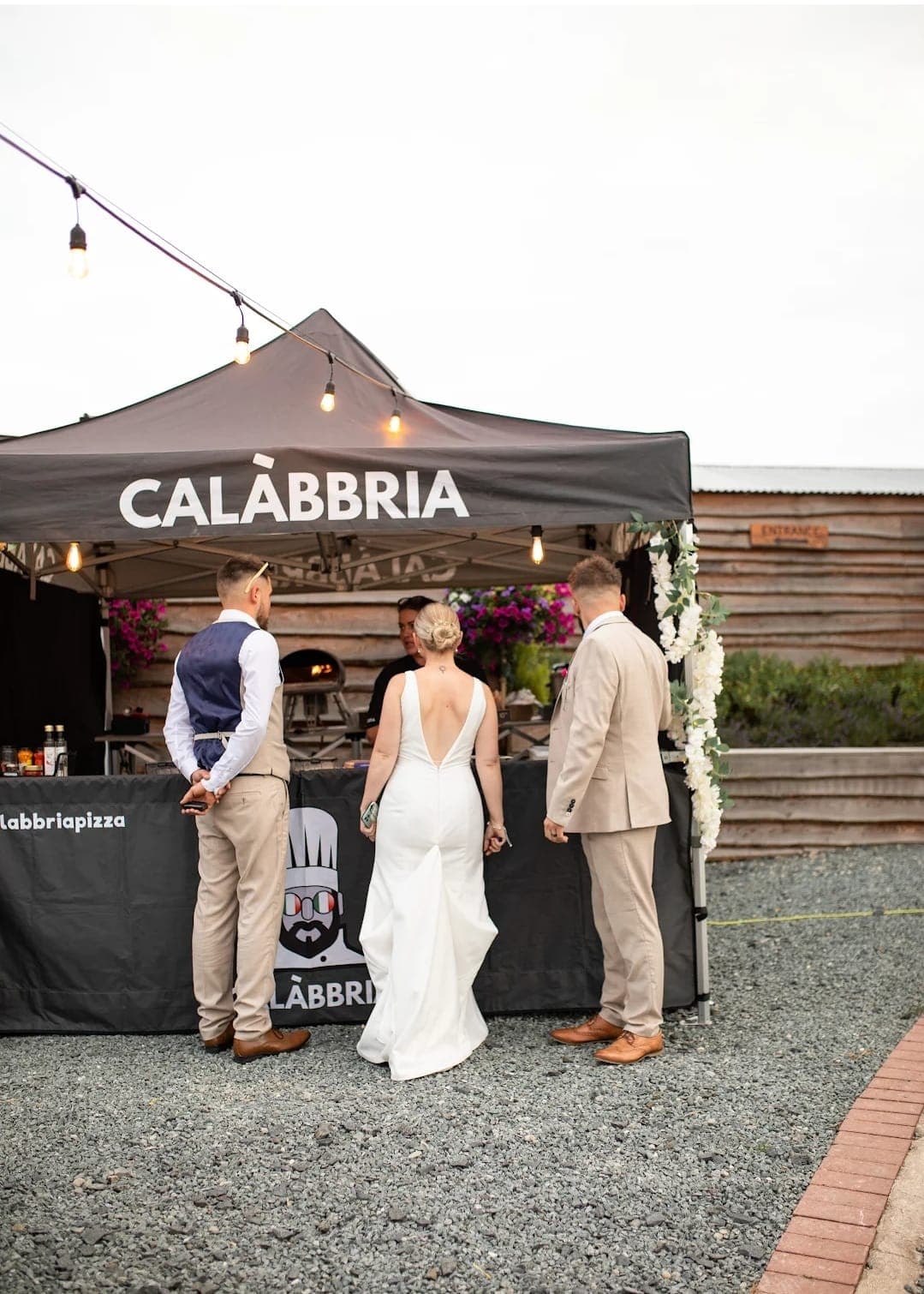 Bride and groom stood at the Calàbbria stall, string lights and floral arch behind them