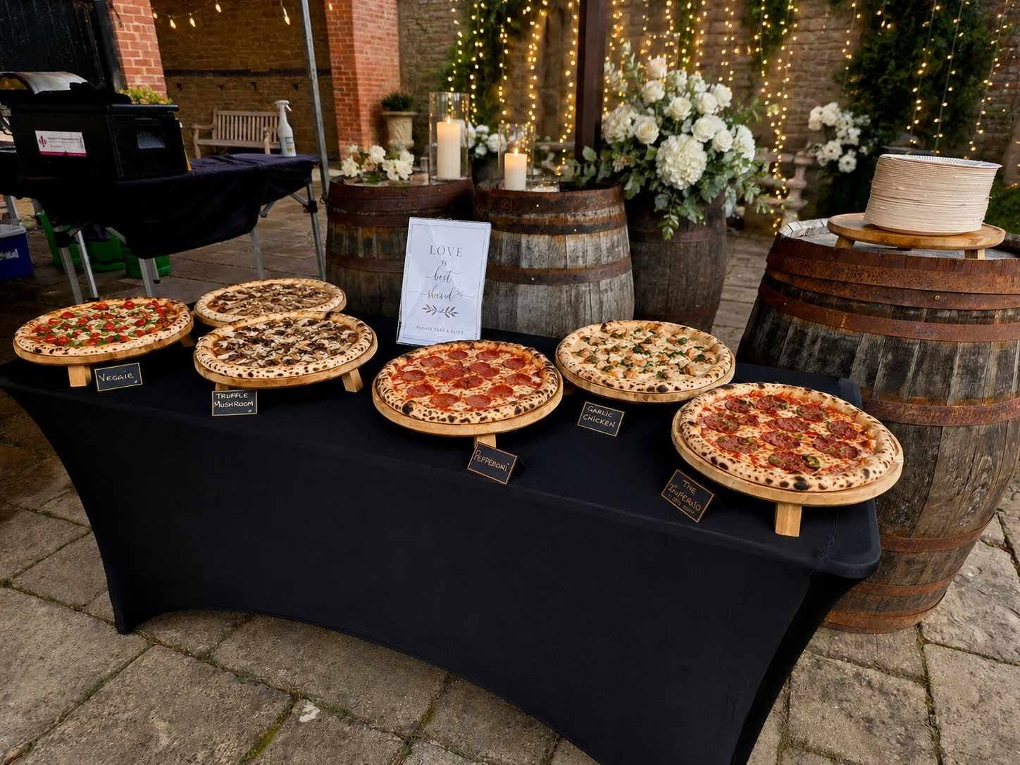 Pizza spread laid out on wine barrels at an evening wedding, fairy lights and white roses behind